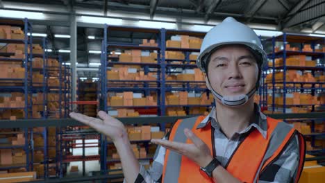 close up of asian male engineer with safety helmet standing in the warehouse with shelves full of delivery goods. smiling and pointing to side recommends about something in the storage