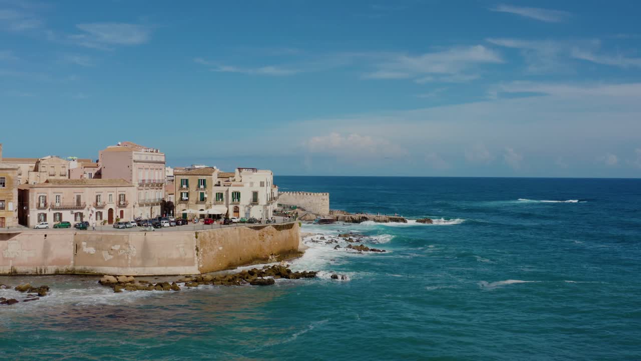 Flying along the fortress wall in Ortigia island with waves crashing on the shore. Aerial view of Ortigia island, Syracuse. Historic town in Sicily.