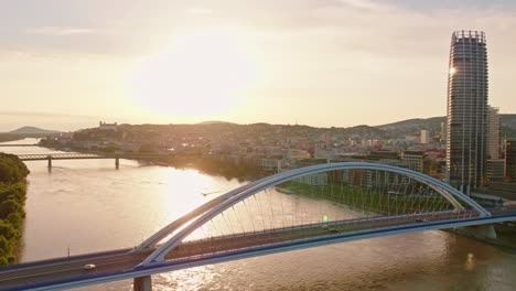 aerial view of bratislava, slovakia, capturing the city's skyline, the iconic bridge over the danube river, and the glowing sunset reflecting on the water