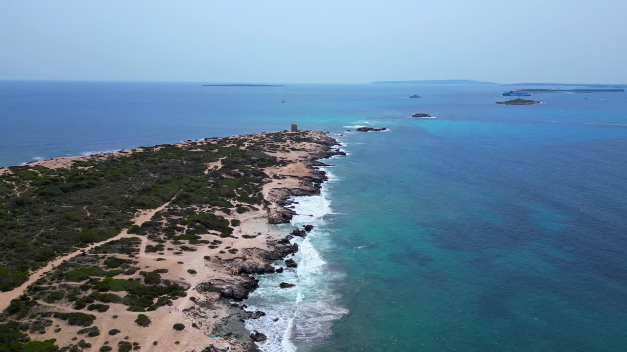 Turquoise waves crashing on the rocky coast of Ses Salines beach with the ancient watchtower. Dramatic aerial view flight drone shot footage from above