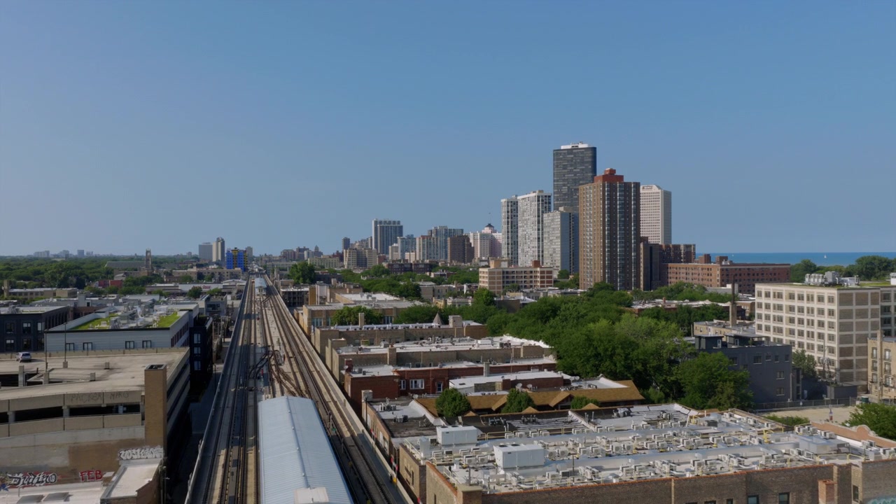 Toma de dron a lo largo de las vías elevadas de la CTA de Chicago hacia un grupo de rascacielos frente al lago bajo un cielo azul despejado