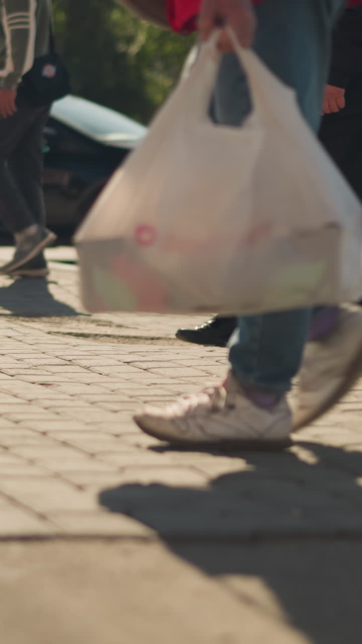Urban crosswalk with pedestrians and cars. Candid daytime scene showing shoppers and commuters stepping across paved sidewalk, sneakers and shopping bags visible, car traffic blurred in background