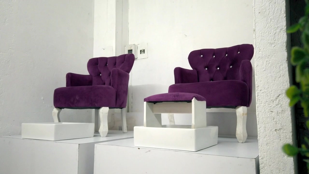 Primer plano panorámico de sillas tapizadas de color púrpura exhibidas sobre plataformas blancas. Respaldos capitoné, reposabrazos curvos, patas de madera y tela similar al terciopelo son visibles en el interior minimalista.