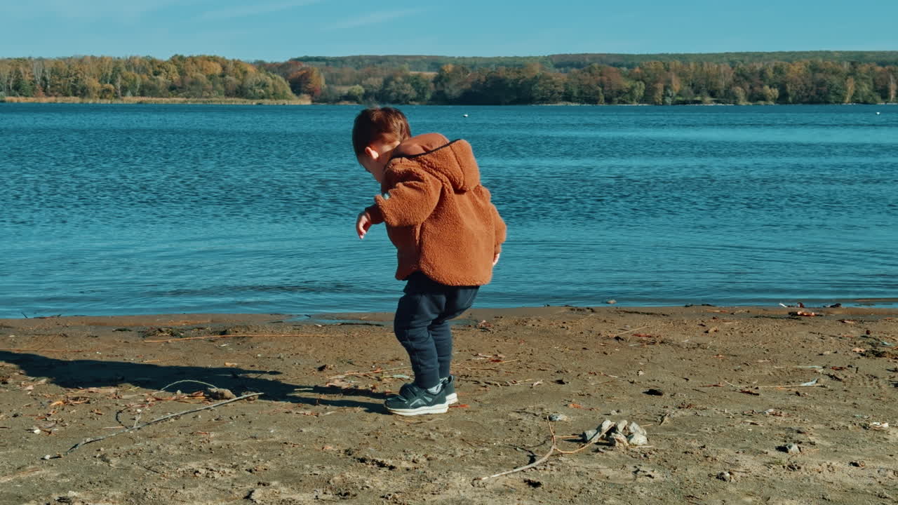 Caucasian male kid throwing stones into water. Baby boy playing on the river bank on sunny day in autumn.