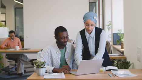 Junger-Arbeiter,-Der-Mit-Laptop-Arbeitet,-Sitzt-An-Seinem-Schreibtisch,-Während-Muslimische-Geschäftsfrau-Mit-Ihm-Spricht-Stand-1