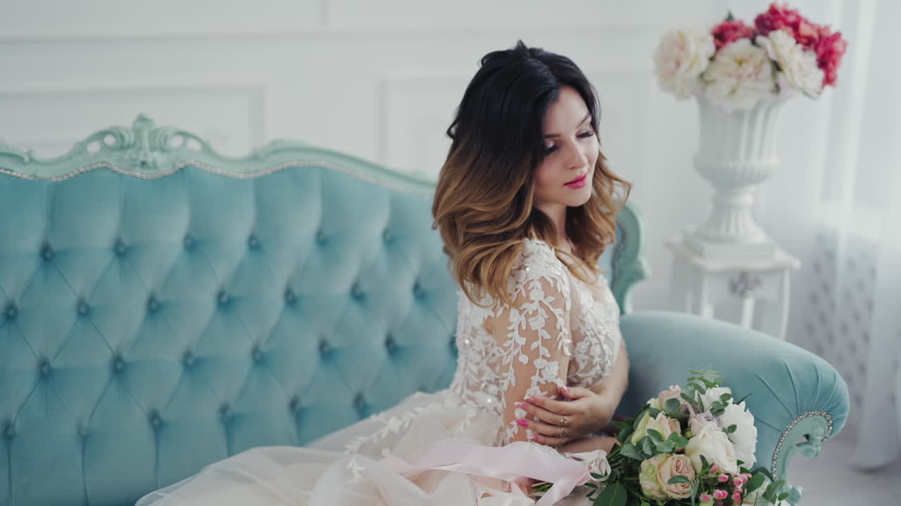 Lovely bride in a nice wedding dress with flowers is sitting on the luxury couch. Beautiful model girl with perfect makeup is posing to camera on a modern studio background.