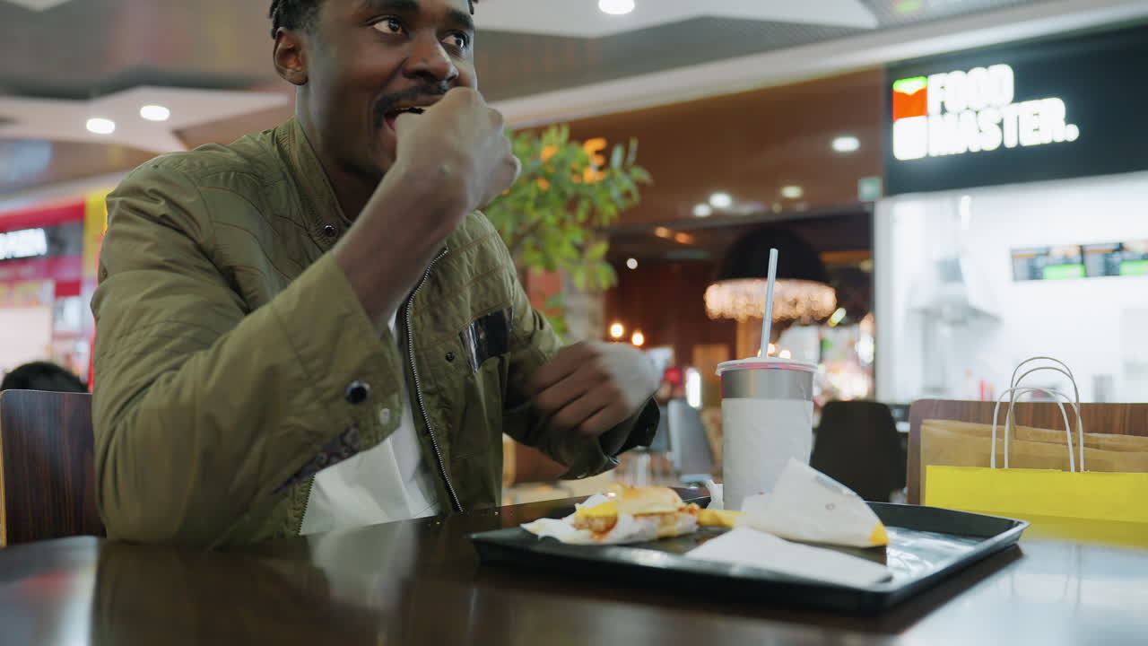 Young freelancer eating sandwich at food court table with drink on tray, wearing green jacket, focused on meal while woman and child pass in blurred background