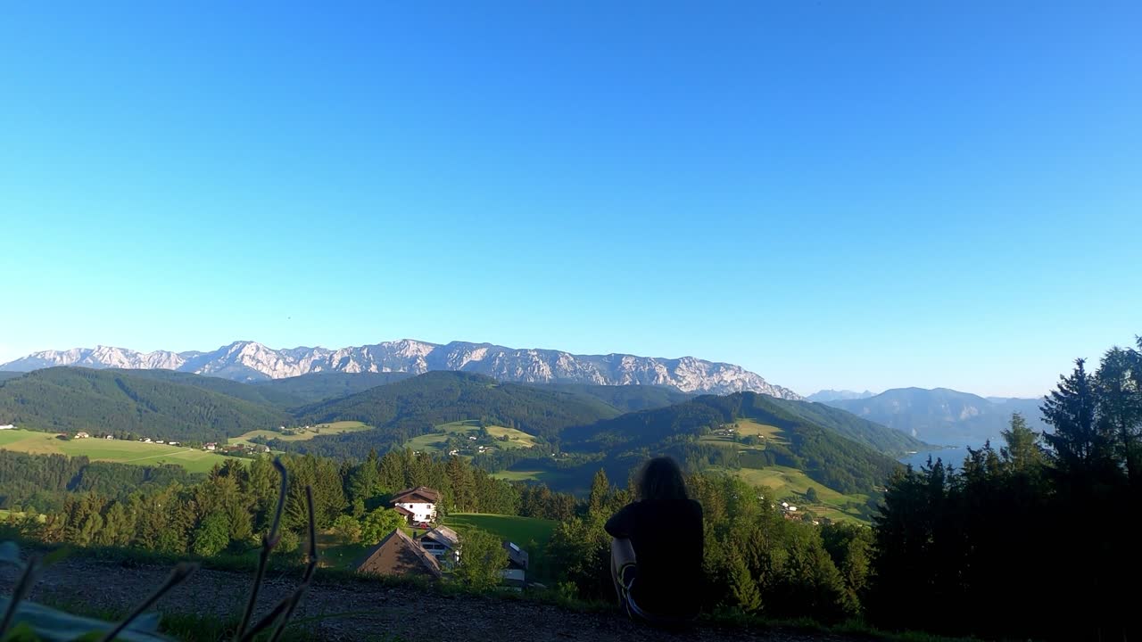Alp dağlarına ve yeşil vadi manzarasına bakan bir yamaçta oturan bir adamın silueti. Doğal panoramik manzaralı, huzurlu bir dış mekan sahnesi.