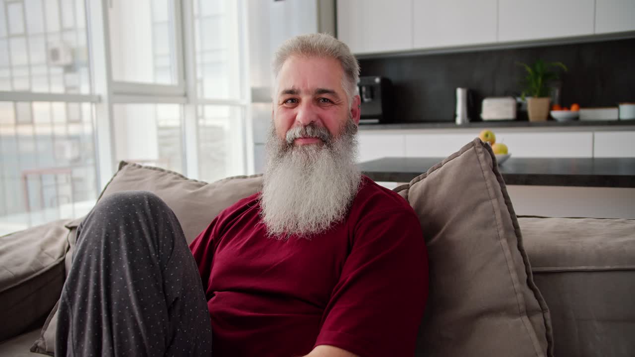 retrato de un hombre mayor feliz con cabello gris y una barba exuberante en una camiseta roja que está sentado en un sofá en un apartamento moderno