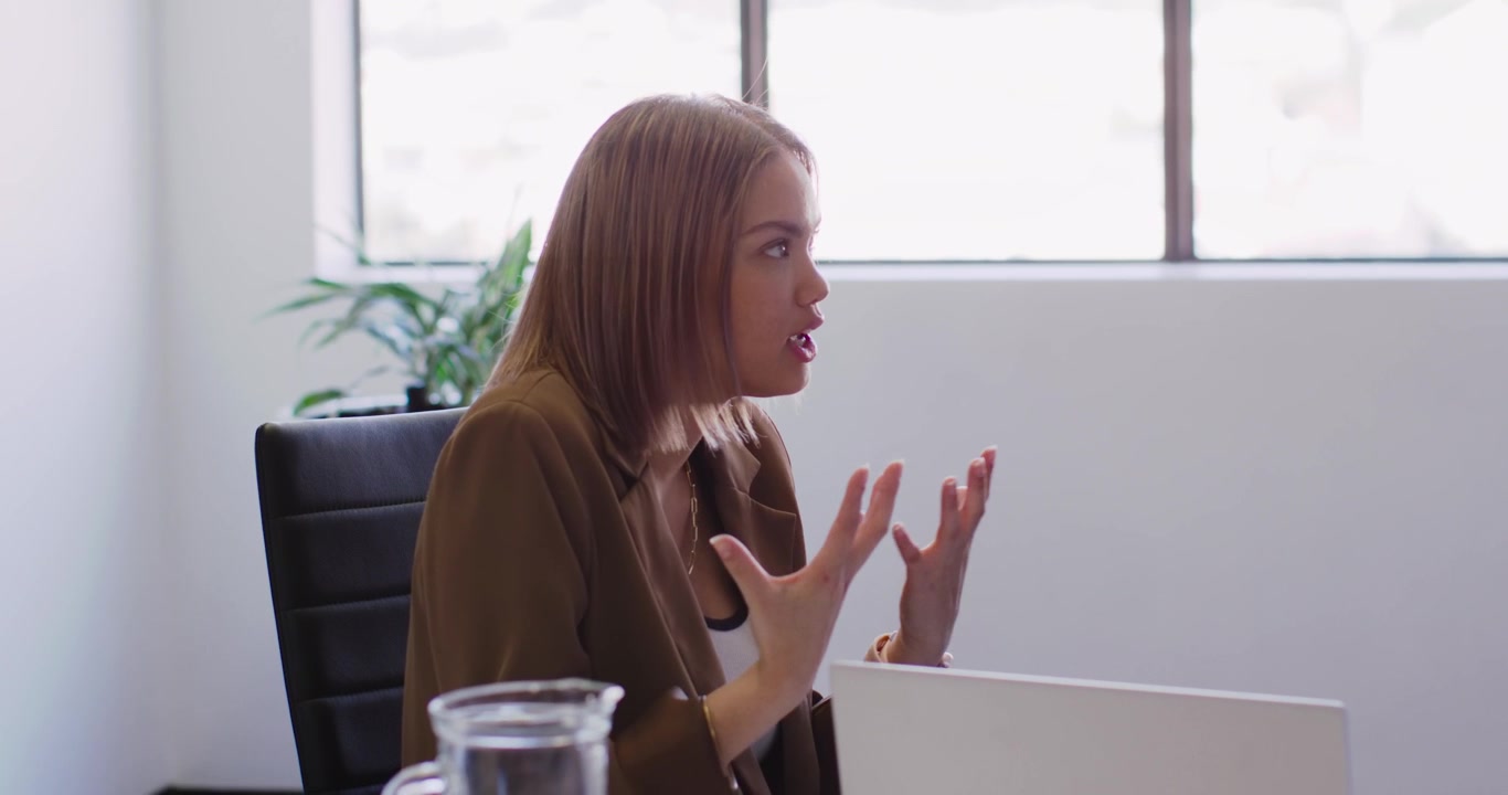 Donna afroamericana in blazer marrone che gesticola per sottolineare un punto, parlando alla scrivania dell'ufficio con un portatile