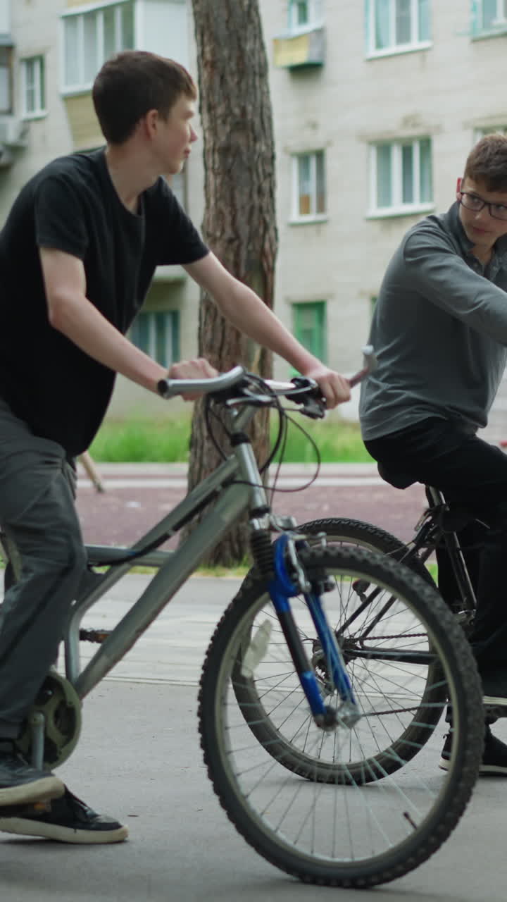 두 소년이 공원에서 자전거를 타고 휴식을 취하는 동안 상호 작용합니다. 한 소년은 회색 과 검은색 바지를 입고 자전거에 앉아서 다른 소년을 바라보고 있습니다.