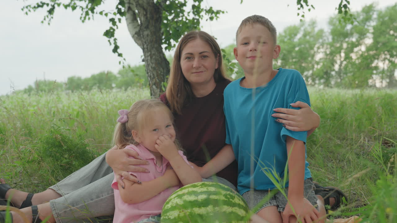 하얀 가족이 나무 아래에서 피크닉을 즐기고 있어요. 엄마는 아들을 안고 있고 어린 딸은 수박과 고리 버들 바구니 옆에 앉아 있네요. 부드러운 여름 햇살, 키 큰 풀, 평온한 시골 분위기, 조용한 관리인이 함께합니다.