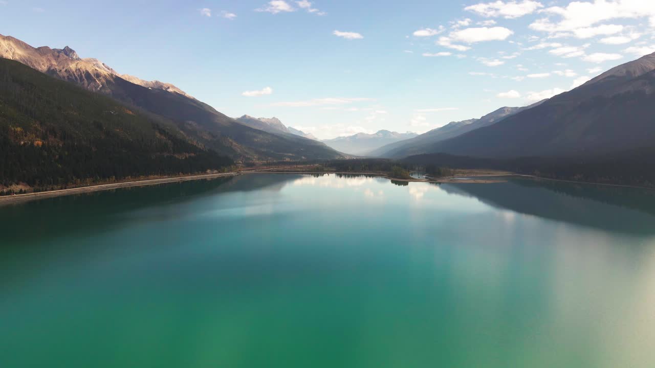 dolly forward aerial wide shot of moose lake in Mount Robson Provincial Park in autumn with yellow trees and mountains in the background and cars driving on the highway with green blue water