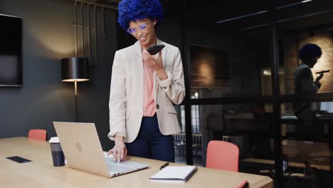 biracial businesswoman with blue afro talking on smartphone and using laptop at desk, slow motion