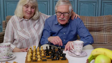 Una-Pareja-De-Ancianos-Jubilados-Hablando-Bebiendo-Té,-Jugando-Al-Ajedrez-En-Un-Moderno-Salón-De-Estar-Juntos