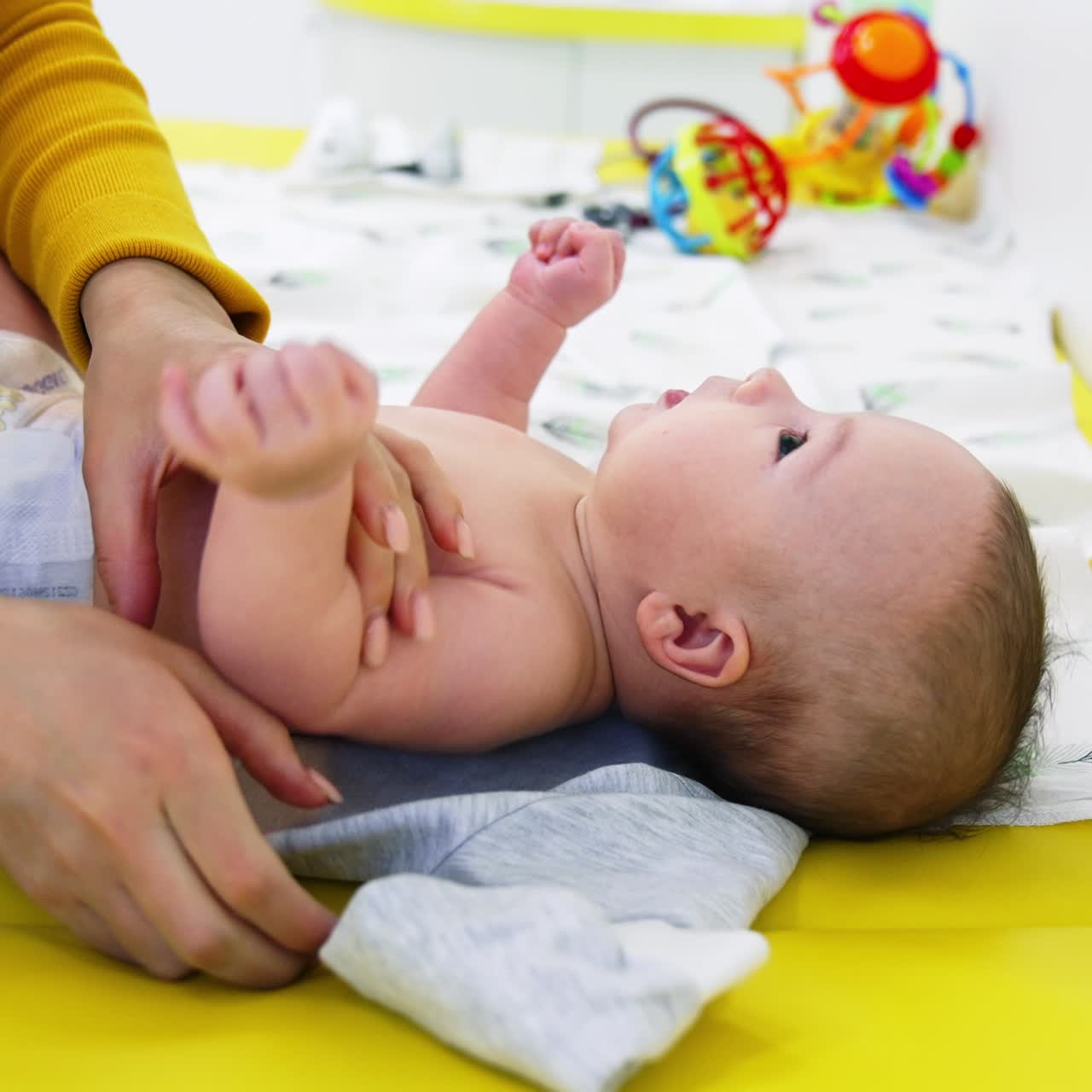 Anne bebeği alıp giysilerin üzerine koyuyor. Bezli bebek yukarı bakıyor. Yan görünüm. Bulanık arka plan