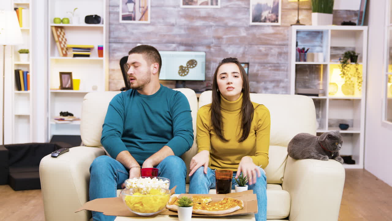 Una coppia e il loro gatto guardano la TV mentre mangiano pizza e snack