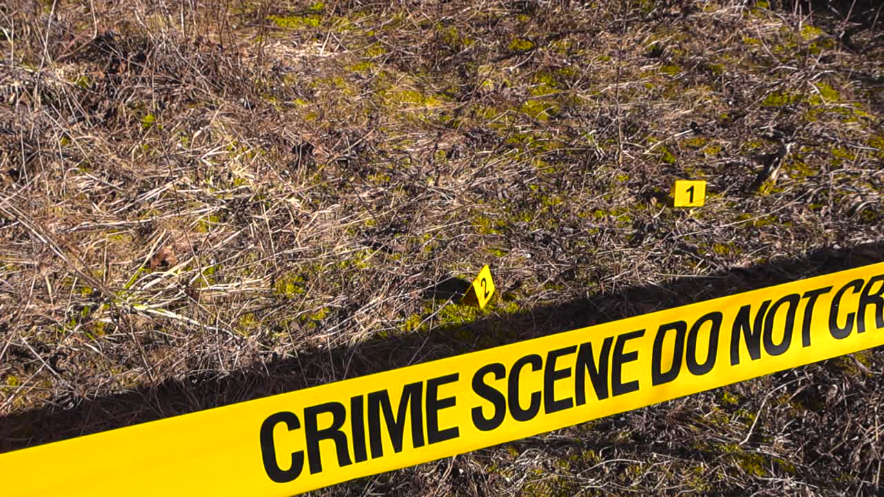 Yellow police tape with the words Crime Scene Do Not Cross cordoning off an outdoor area with numbered evidence markers placed on the dry grass, indicating a forensic investigation