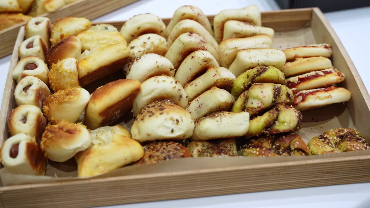 Primo piano di un assortimento di mini pasticcini salati appena sfornati, inclusi involtini ripieni di formaggio, bocconcini con sesamo e pezzi di impasto aromatizzato, disposti in un vassoio di legno per l'esposizione in un buffet catering.