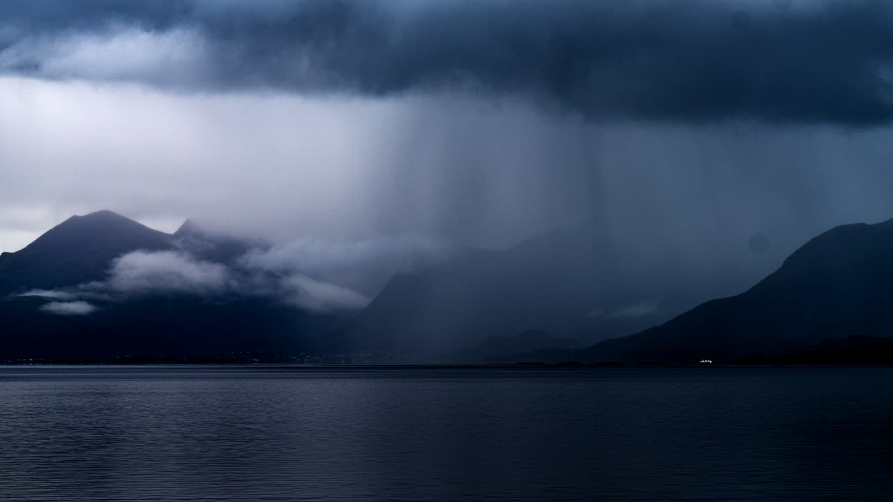 ノルウェー、ヴェステローレン山脈とフィヨルドの上空を覆う嵐の雲と舞い踊る雨柱のタイムラプス映像。背景素材。