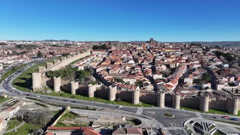 Seitlicher-Flug-In-Der-Weltkulturerbe-Stadt-Avila-Mit-Blick-Auf-Die-Mittelalterliche-Stadtmauer-Mit-Ihren-Innenhäusern-Und-Ihren-Grünflächen-Mit-Einer-Straße,-Auf-Der-Fahrzeuge-An-Einem-Wintertag-Mit-Blauem-Himmel-In-Spanien-Zirkulieren