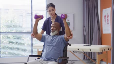 una fisioterapeuta femenina diversa y un paciente masculino anciano sosteniendo pesas en una sesión de fisioterapia.