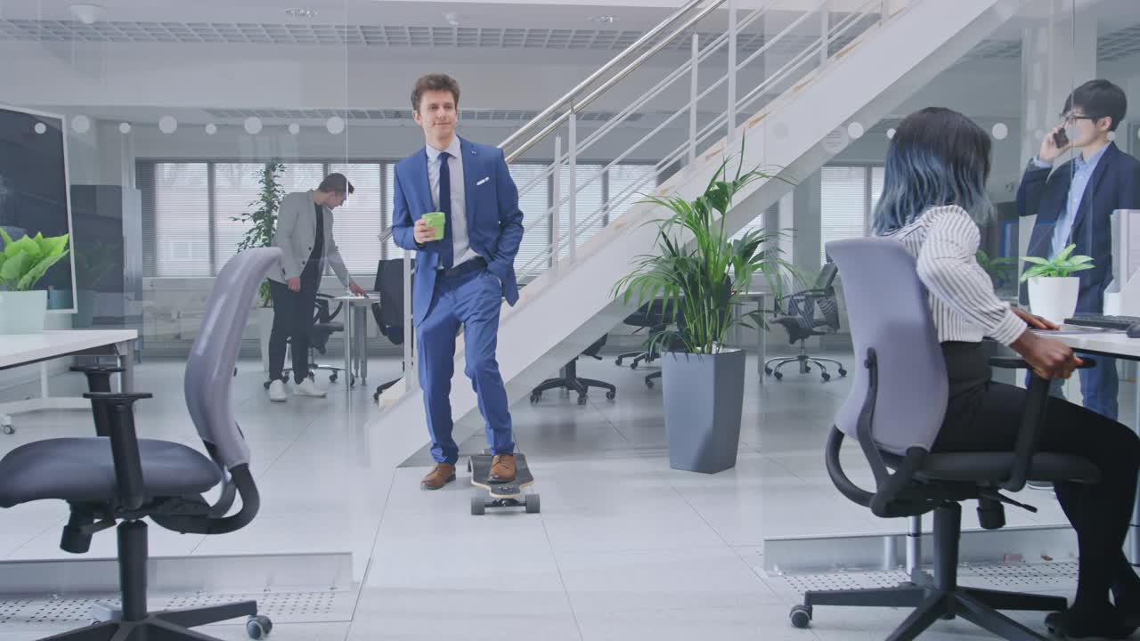 Young Happy Handsome Manager Wearing a Suit and Tie Rides a Longboard. He's Holding a Cup of Coffee. Diverse and Motivated Business People Work on Computers in Modern Open Office.