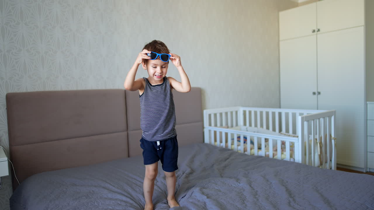 Little Caucasian toddler wearing sunglasses jumps on the bed. Happy baby boy playing at home.