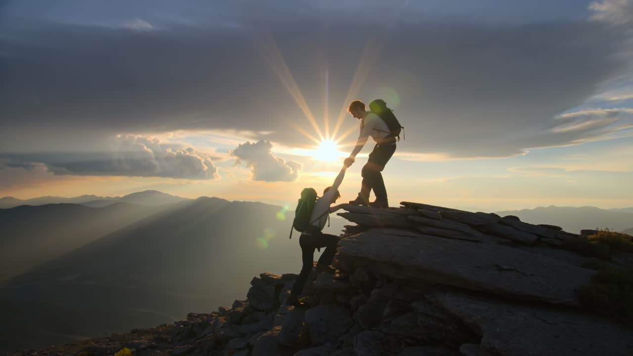 Hiker reaching out to help a partner climb a steep rocky mountain peak at sunrise