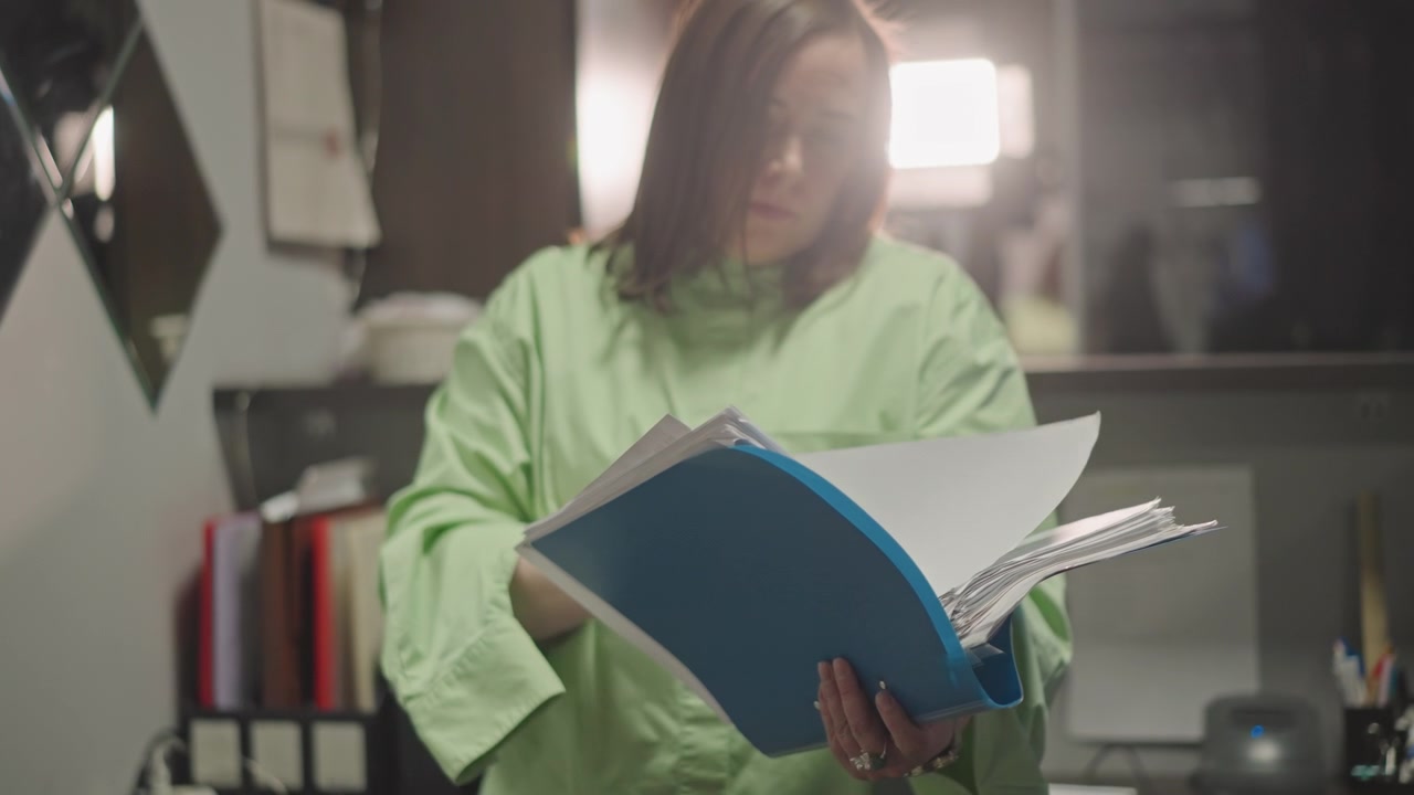 Insegnante di musica che esamina un raccoglitore blu, con controluce drammatico e un leggero bagliore dell'obiettivo; scaffali per documenti e penne visibili. Sfoglia le pagine dello spartito con espressione concentrata, durante la preparazione tardiva per le prove, alla scrivania dello studio, focalizzato.