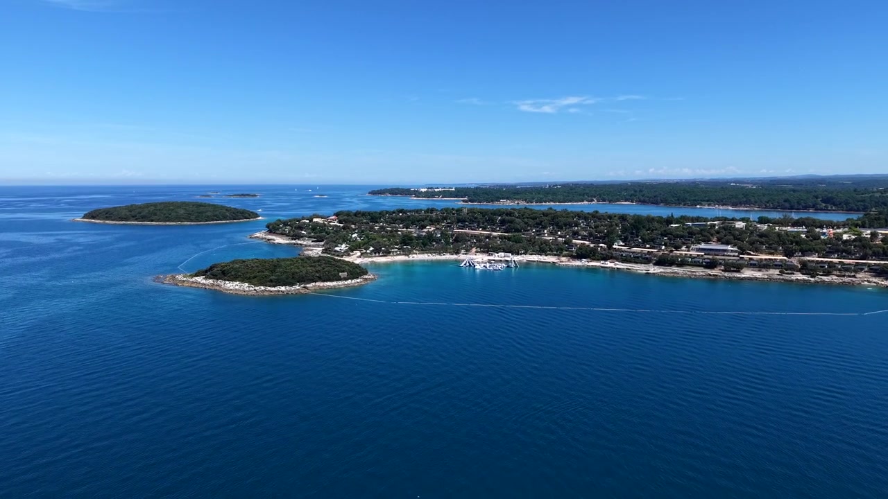 Wyspy porośnięte sosnami rozsiane po czystych wodach Adriatyku u wybrzeży półwyspu Istria, z nadmorskim ośrodkiem kempingowym i zalesioną linią brzegową rozciągającą się pod jasnym letnim niebem. Chorwacja. Szerokie ujęcie z drona wznoszącego się.
