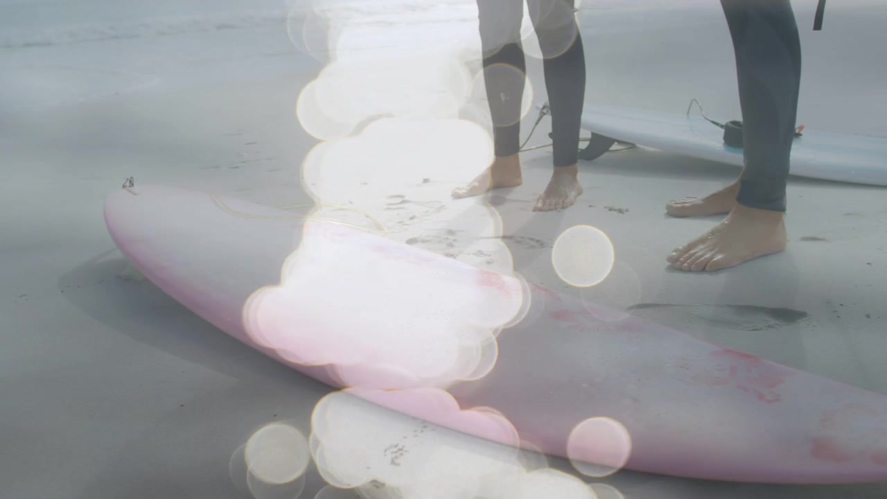 Uomo e donna in piedi sulla sabbia, con effetto bokeh che rivela una tavola rosa e i leash, mentre si preparano per il surf.