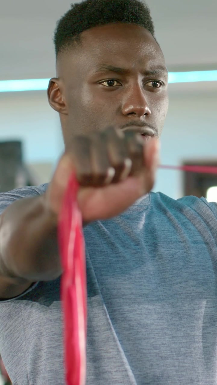 Vertical video: Gripping red band chest-high, fit man in blue tee pulling arms in gym for strength