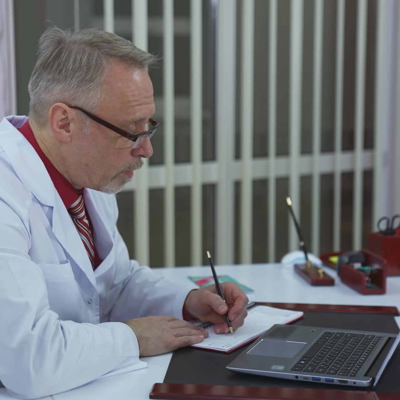 Doctor sitting at desk in medical center during telemedicine. Medical specialist in white coat and glasses making online video call on a laptop.
