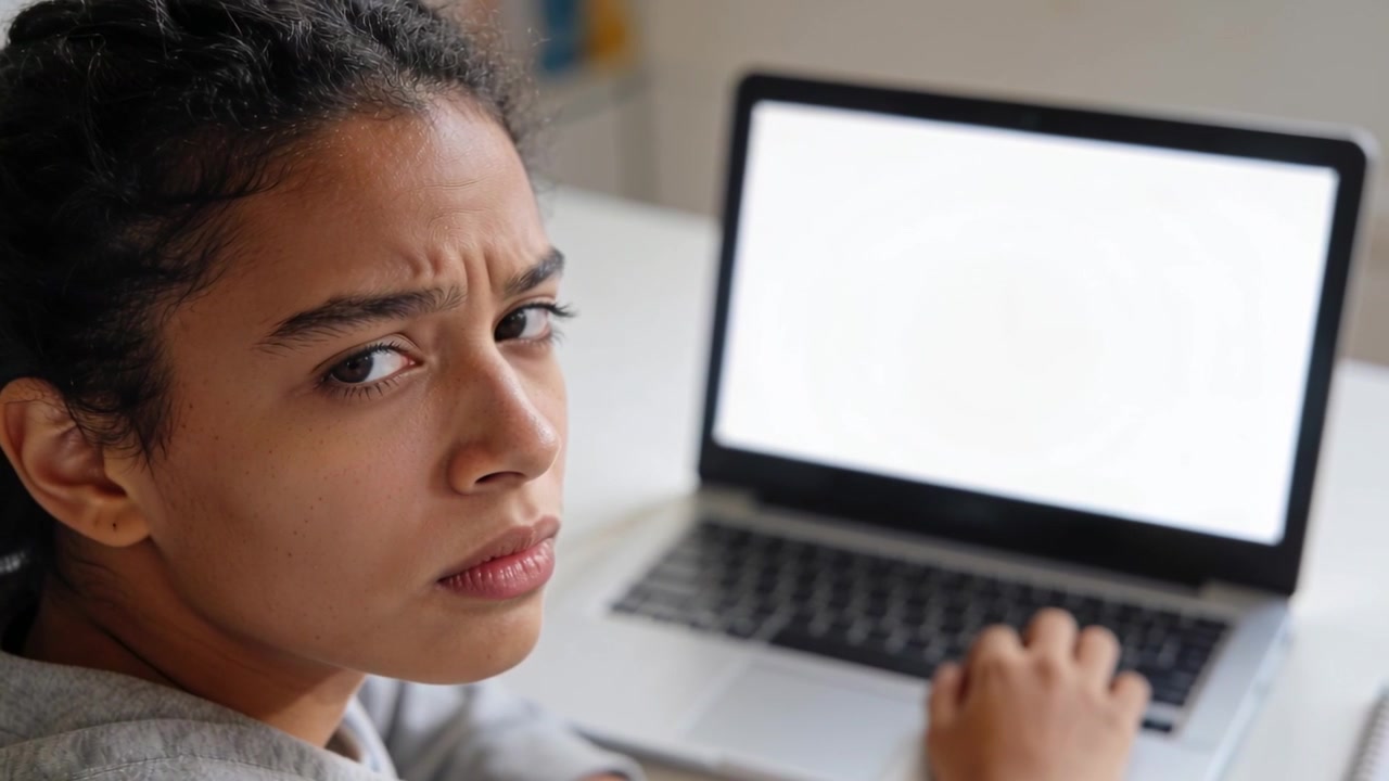Eine Frau im grauen Kapuzenpullover dreht sich am Schreibtisch um, die Hand auf der Laptoptastatur, irritiert von einem leeren Bildschirm.