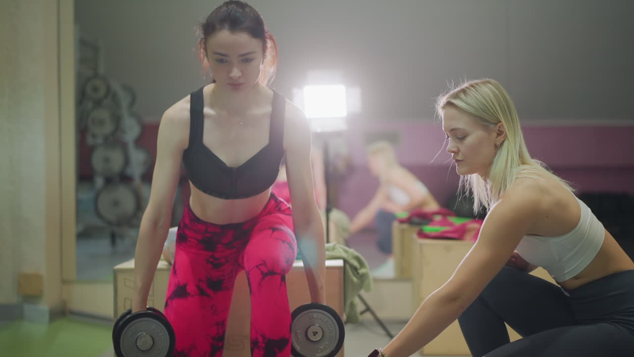 Mulher levantando halteres ao lado da treinadora em estúdio de academia, treinadora ajoelhada ajustando o posicionamento dos pés, leggings rosa vibrante, roupa esportiva, caixa de madeira, supervisão próxima, ritmo deliberado e focado.