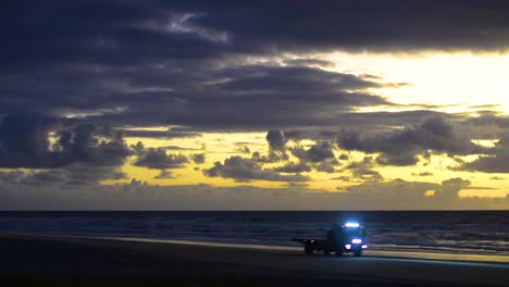 dramatic and cinematic blue and yellow sunrise sunset over a calm pacific ocean beach scene with a silhouetted brightly-lit flatbed truck driving along the beach