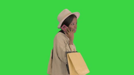 niña afroamericana con ropa de punto y sombrero de gran tamaño hablando en su teléfono mientras camina con bolsas de compras en una pantalla verde, llave de croma