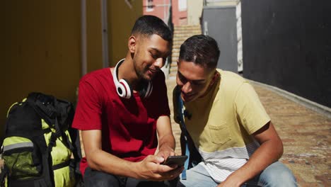 dos amigos de raza mixta sentados y usando un teléfono inteligente en la calle