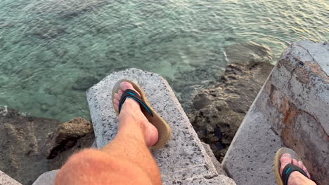 the camera tilts up from the filmer’s legs gliding over calm, glassy water toward the golden-orange glow of the sun just set behind a distant mountain ridge, under soft clouds and a peaceful sky