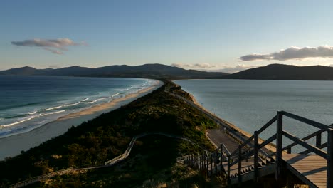 Stunning-timelapse-of-the-neck-isthmus-on-bruny-island-during-orange-sunset,-tasmania,-australia,-coastal-beach-waves-rolling-in-on-white-sand-with-lush-green-forest-on-clear-sky-evening