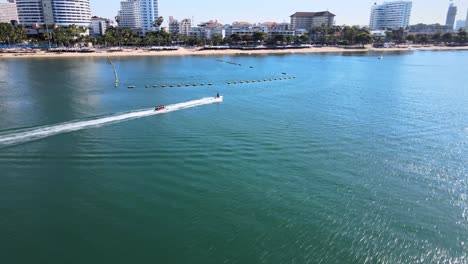 4K-Drohnenaufnahmen-Eines-Jetskis,-Der-Ein-Boot-Durch-Meereswellen-Zu-Den-Wunderschönen-Ufern-Von-Pattaya-An-Der-östlichen-Golfküste-Thailands-Zieht