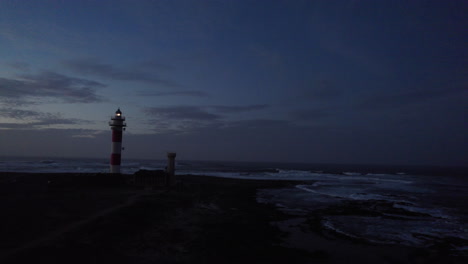 aerial footage of a lonely lighthouse at the rocky ocean shore with yellow light on top at dawn with waves crashing all around and dark blue sky in back