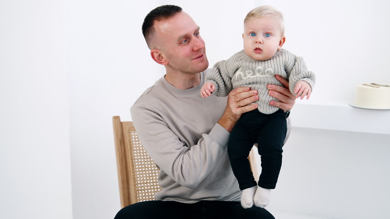 Happy Caucasian man in grey sweater holding his little baby in grey sweater too. Father and son portrait at white backdrop.