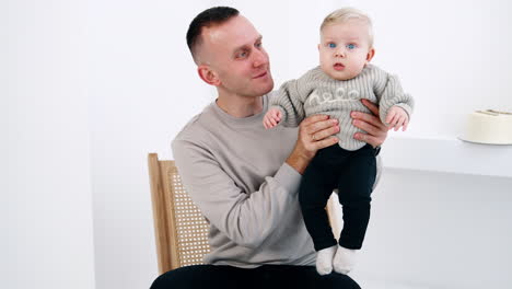 Happy Caucasian man in grey sweater holding his little baby in grey sweater too. Father and son portrait at white backdrop.