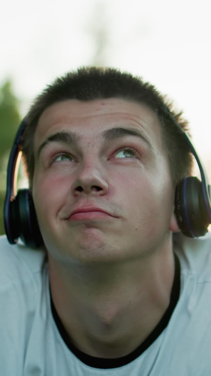 Close up of music artist lying on grass wearing headphones nodding to music while using phone, looking upward thoughtfully, with soft bokeh effect from trees