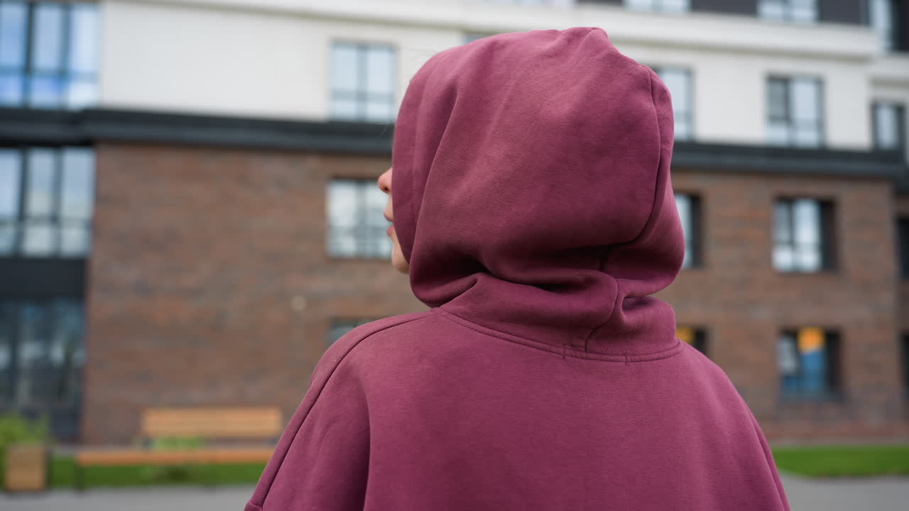 Back shoulder view of young woman in maroon hoodie with covered hair strolling along paved path toward gym facade past wooden playhouses and spring foliage near urban courtyard wearing white sneakers