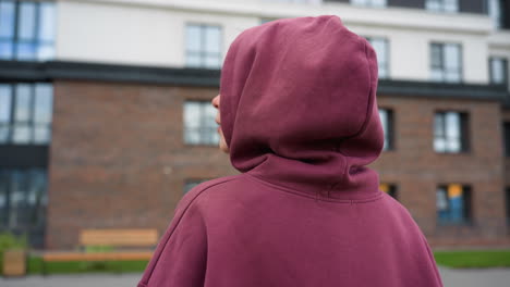 back shoulder view of young woman in maroon hoodie with covered hair strolling along paved path toward gym facade past wooden playhouses and spring foliage near urban courtyard wearing white sneakers