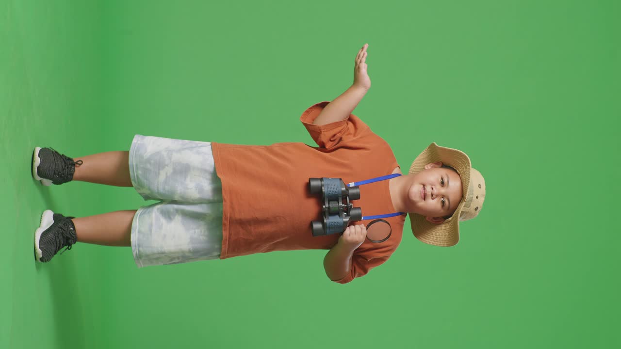 Asian Tourist Boy With A Hat And Binoculars Holding The Magnifying Glass And Pointing To Side While Standing On Green Screen Background. Boy Researcher Examines Something, Travel Adventure, Full Body