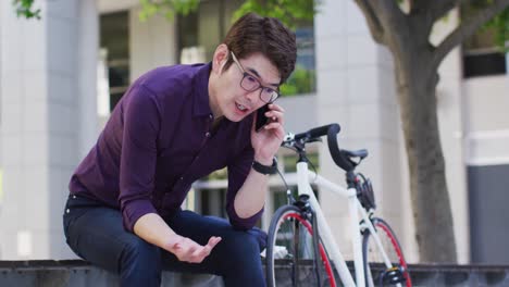 Hombre-Asiático-Hablando-Por-Teléfono-Inteligente-Mientras-Está-Sentado-Cerca-Del-Parque-Corporativo